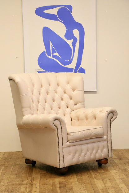 Cream leather Chesterfield wing armchair with deep-buttoned high back, rolled arms, brass studs and bun feet